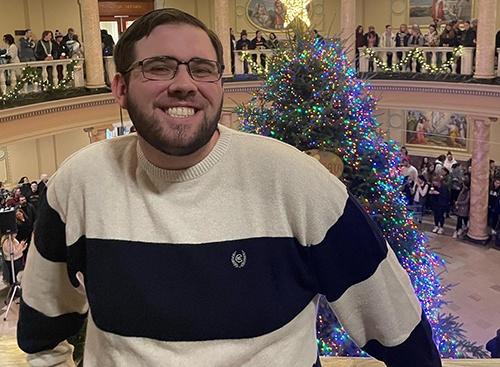 Man smiling in a blue and white striped shirt with a christmas tree lit in the background.