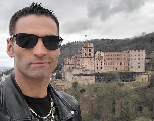 Man in sunglasses and a black leather jacket posing in front of a historic castle on a cloudy day.