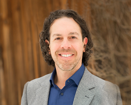 Alumnus Michael E. Bugno, smiling, wearing a gray blazer and blue shirt, with a wooden background.