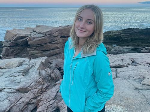 Young woman smiling wearing a blue jacket with rocks and the ocean behind her.