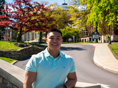 Young man in a light blue collared shirt smiling outside with trees in the background.