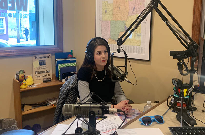 Woman sitting at a radio station desk, wearing headphones and speaking into a microphone.