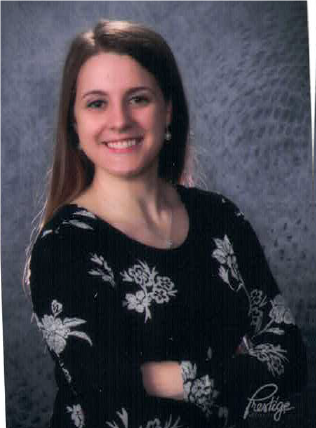 A young woman in a dark dress floral pattern dress, confidently smiling against a grey background.