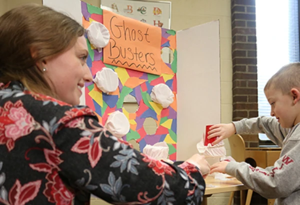 Woman smiling wearing a printed blouse helping a young child in a classroom setting.