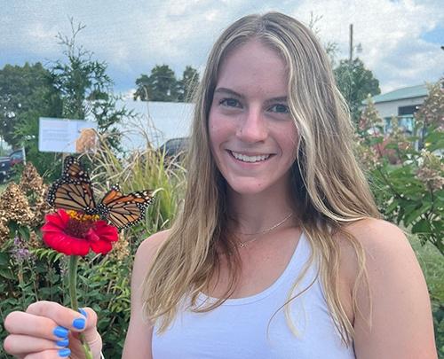 Young woman smiling wearing a white tank top holding a flower with a butterfly on it.