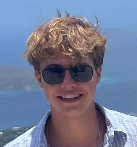 Young man smiling wearing a collared shirt and black sunglasses with a body of water in the background.