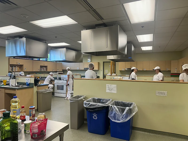 kitchen area with ingredients on the table and students in white scrubs and hats talking to eachother