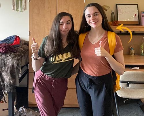 Two young women smiling holding a thumbs up in a dorm room.