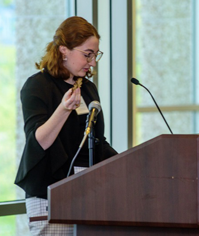 A woman with red hair and glasses, wearing a black blazer over a white outfit, speaking into a microphone at a podium.