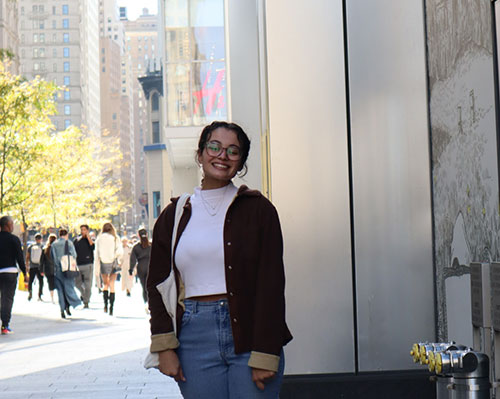 Young woman smiling wearings jeans, a white top, and a black sweater with buildings and people in the background.