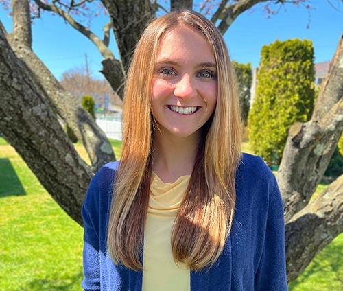 A woman with long straight blonde hair and blue cardigan, smiling in an outdoor setting with trees and a clear sky in the background.